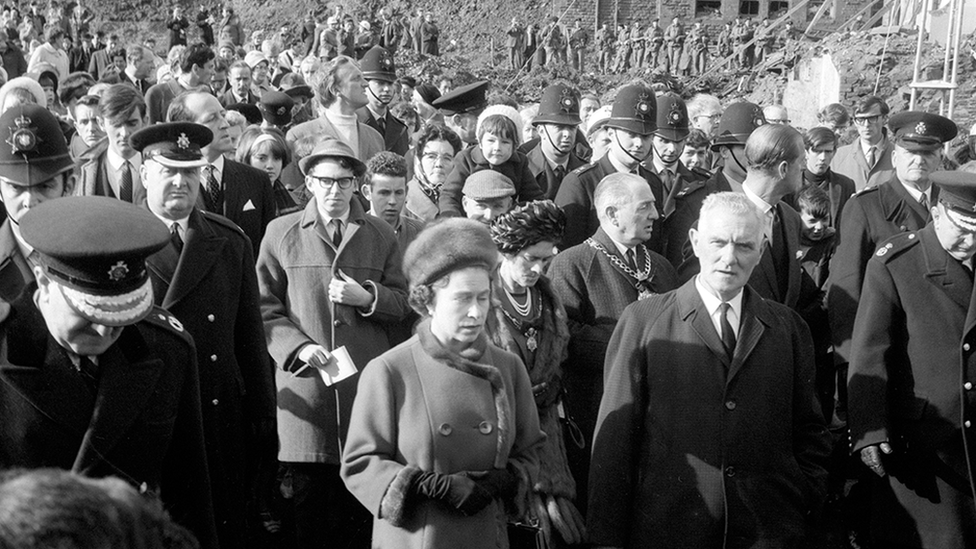 A black and white photo shows the late Queen Elizabeth walking at the front of a large group of people. She wears a fur hat and coat with fur lining.