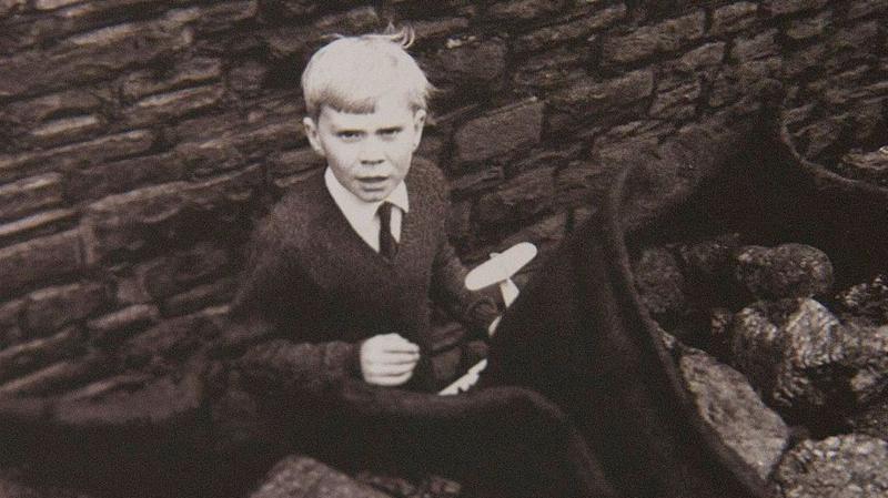 A young boy with white hair, and wearing a jumper, white shirt and black tie, plays in the street behind sacks of coal. He is sat down, with a brick wall behind him.
