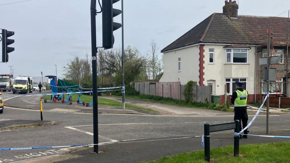 The house where the murder took place, pictured from across the street behind blue and white police cordon tape. The white end-of-terrace property has a wooden fence bordering the side of the house and stretching into the rear garden. There is a police officer in a yellow hi-vis vest standing on the pavement, guarding the cordon.
