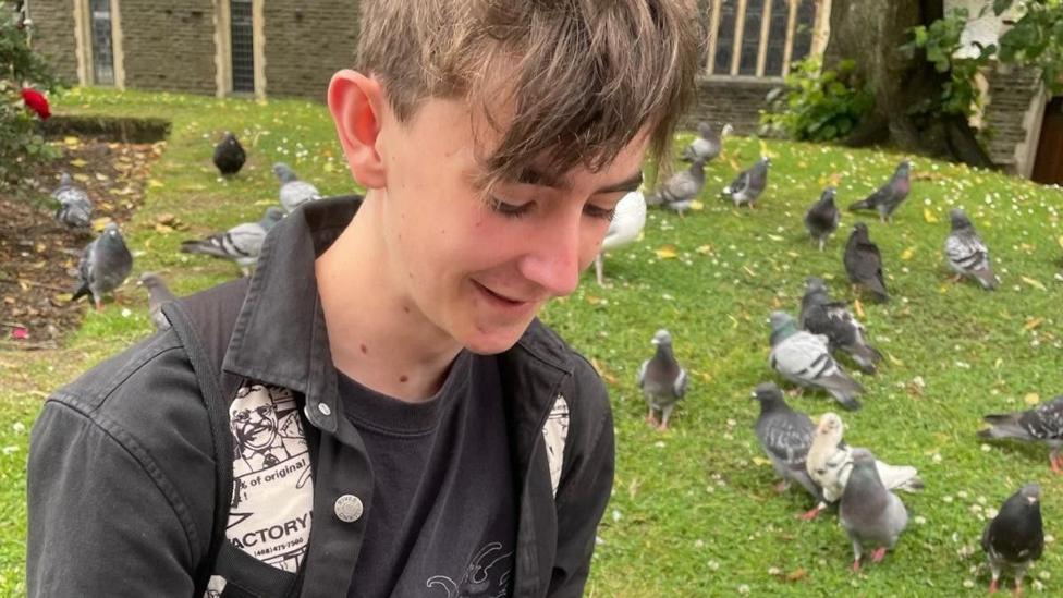 Jamie Collins wearing a black denim jacket and backpack with black and white comics on the shoulder straps. He is sitting in a green park area surrounded by pigeons, with a brick church visible in the background.