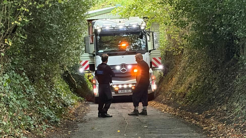 A police officer and a man talking in front of a lorry, which is pictured trying to transport the caravan up a narrow road, scraping trees and branches.