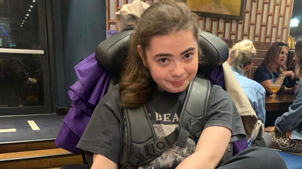 Romi sitting in a supportive wheelchair in a restaurant or café setting, wearing a dark graphic T-shirt. The image is taken indoors, with other diners seated at tables in the background and large windows visible to one side.