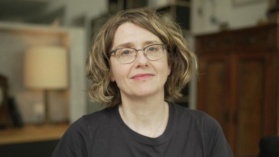 Close-up portrait of Gael with short wavy hair and glasses, wearing a dark top and sitting indoors. A lamp, shelving and wooden furniture can be seen in the softly focused background.