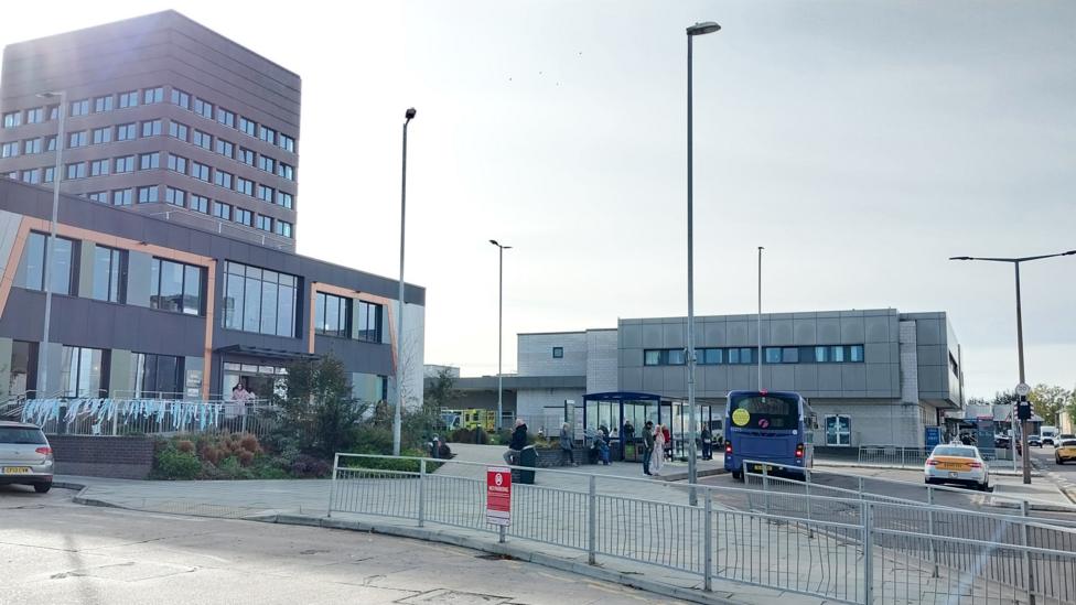 Exterior views of several hospital buildings. To the left is a taller, high rise type building. In the foreground is a road and a pedestrian barrier. Cars and a bus are visible alongside people waiting at a bus stop.
