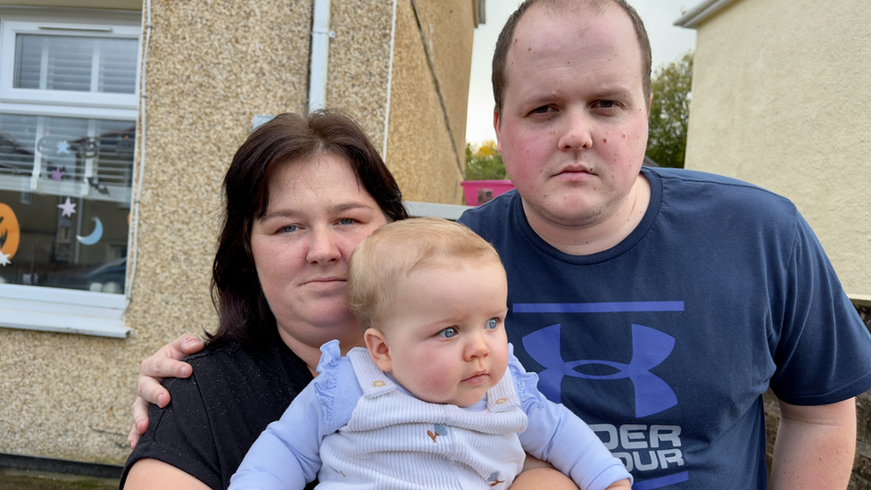 Rhiannon and Adam stand outside a house, Adam with his hand around Rhiannon's shoulders. Rhiannon has long dark hair, Adam has short dark hair and is clean shaven. Rhiannon holds baby Summer-Gracie in her arms, she is wearing a purple outfit and has short blonde hair.