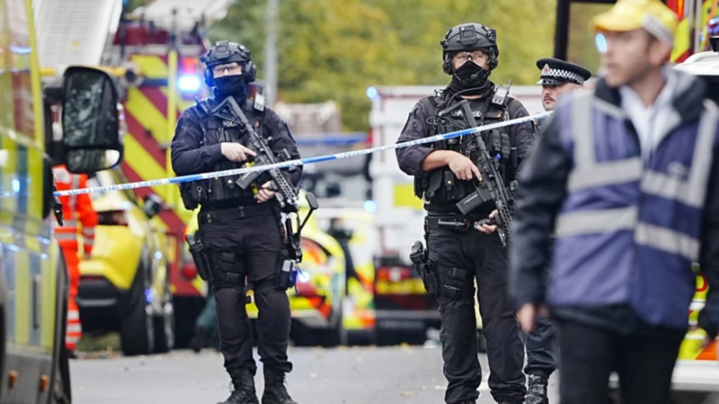 Armed police officers in protective tactical gear stand behind a police cordon at an emergency incident scene after an attack at Heaton Park Hebrew Congregation Synagogue. Several emergency vehicles with flashing lights are visible in the background, while a blurred person wearing a high-visibility vest stands in the foreground. The street is partially closed off with police tape, and trees with autumn foliage can be seen in the distance.