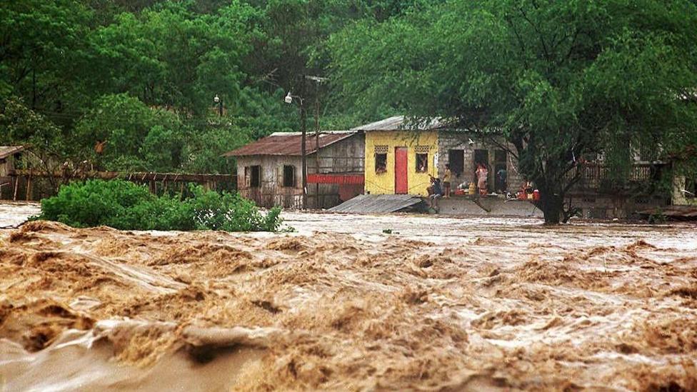 Torrent of flood waters raging through a settlement with people stranded on the roof of their building surrounded by flood water
