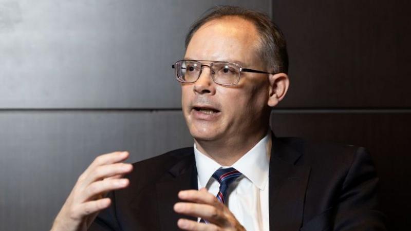 Tim Sweeney looks to the side as he is interviewed. His hands make motions as he answers a question. The Epic Games boss wears a white shirt, blue and red striped tie and black suit jacket.