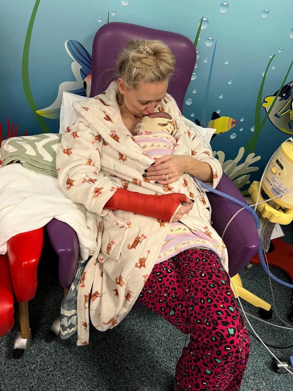 Jo sits in a purple hospital chair holding newborn baby Phoebe wrapped in a pastel knitted blanket. The baby is wearing a respiratory support mask secured with a soft headband and is connected to medical tubing. Jo is wearing a patterned dressing gown, bright pink trousers and a red arm cast. The hospital room features a colourful underwater-themed wall mural, with medical equipment and cables visible beside the chair.