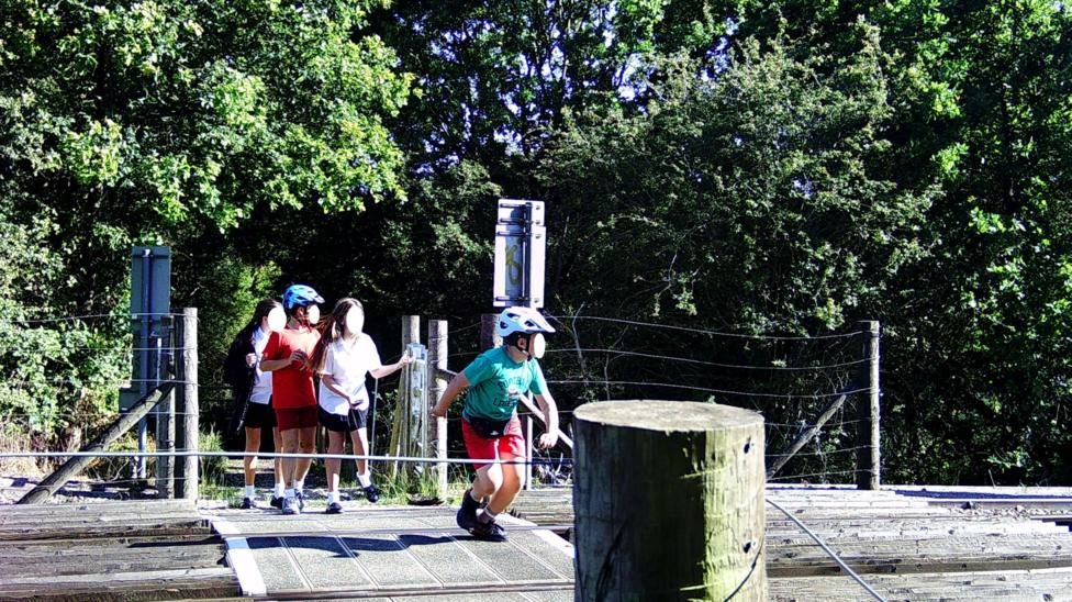 Four children on a railway level crossing surrounded by trees. A boy in a white cycling helmet, green T-shirt and red shorts runs across the tracks in the foreground, while three others stand by the gate behind him. Their faces are blurred.
