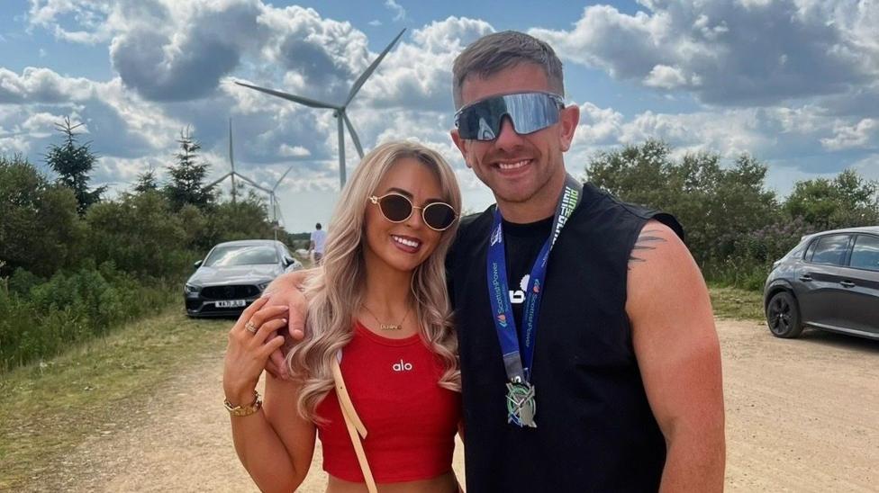 A couple standing outside on a mostly sunny day, with their arms around each other. Both are wearing sunglasses. Denby McLaren is wearing a red top and Craig Anderson is a sleeveless black top on. Two cars are visible behind them, as is a large windmill.