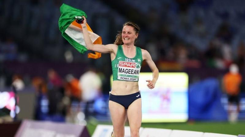 A woman celebrates on an athletic track, she waves an Irish flag