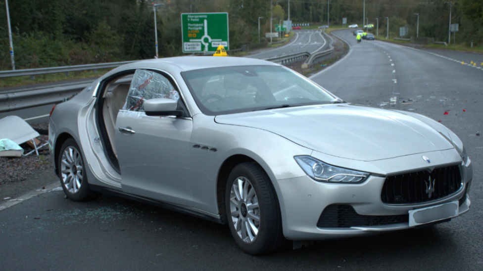 Front view of a grey Maserati car in the middle of a lane on a dual carriageway. The car is visibly damaged with the air bags blown up and windows smashed. There is debris around the car on the road. Police officers can be seen in the distant background.