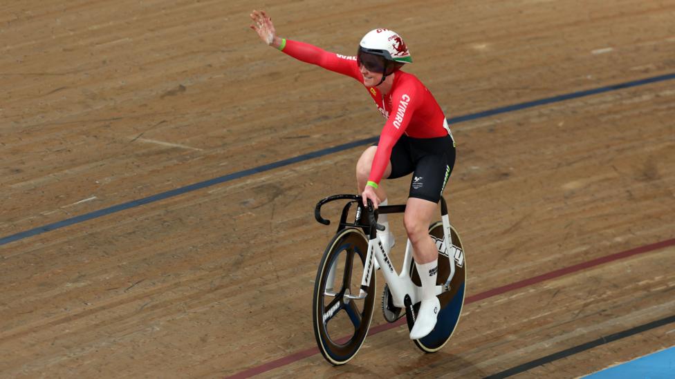 Emma Finucane of Team Wales in red and black waves while riding a balck and white bike a velodrome track