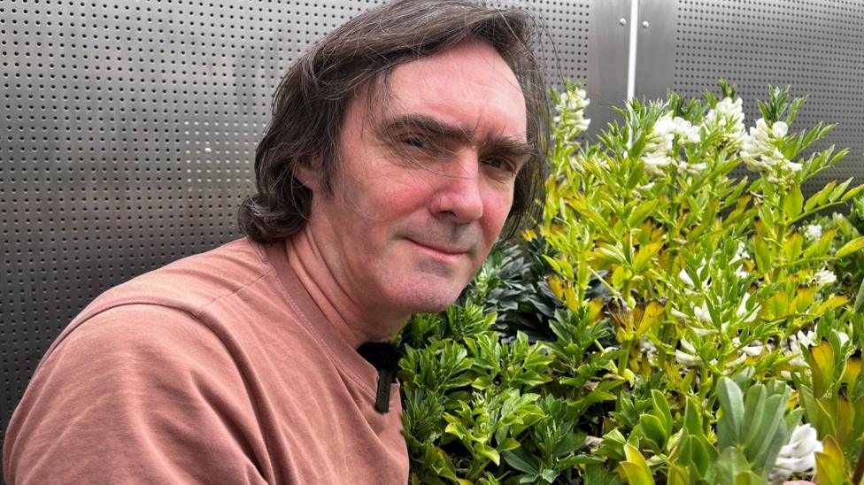 A man in a red t-shirt with brown hair in front of some fava bean plants.