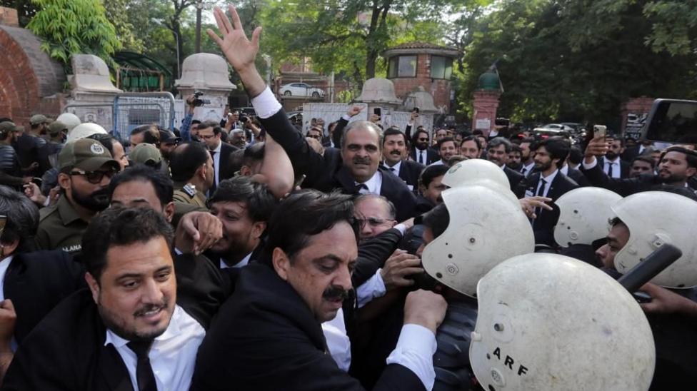 Dozens of men in dark suits scuffle with men wearing white protective helmets in a street in Khyber Pakhtunkhwa