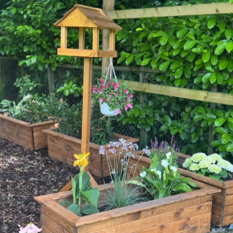 Wooden planter in the foreground containing flowering and leafy plants, with a wooden bird-table-style structure mounted in the centre. A hanging basket of pink flowers is suspended from the structure. Additional raised wooden planters line a fenced garden behind it, set on a bark-covered surface. A small garden ornament stands beside the planter, and dense green hedging forms the background.