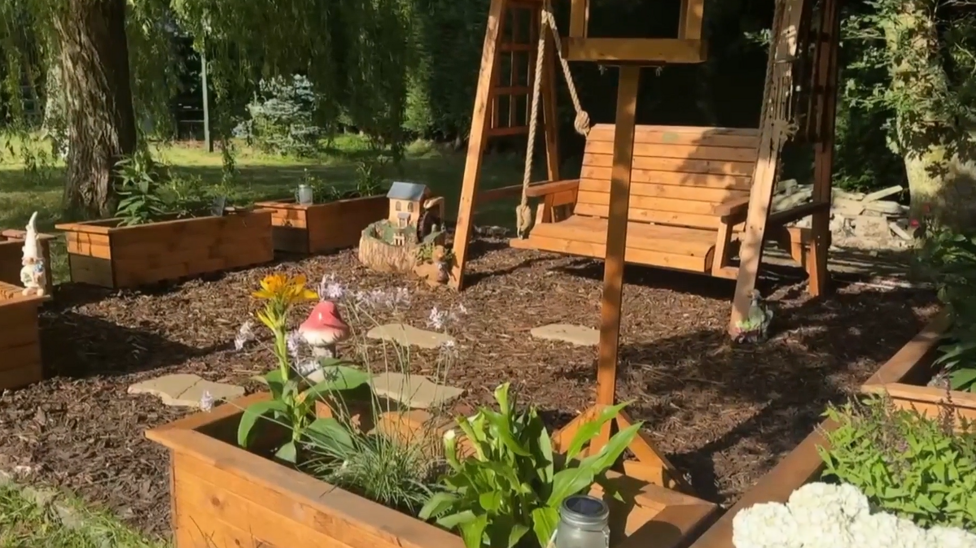 Wooden garden swing suspended from a timber frame in the foreground, surrounded by raised wooden planters filled with plants and flowers. Stepping stones run across a bark-covered surface beneath the swing. A small decorative house and garden ornaments are positioned among the planters. Mature trees and dense greenery form the background.