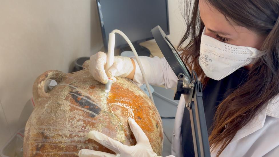 A close up view of an amphora in the laboratory on the ship. The handle on the side of the pot is poking out to the left. A woman with long brown hair, wearing a white lab coat and a face mask, is working on the surface of the amphora with a small electric tool. It's white and is about 10cm long with a light on the end, she is holding it in her right hand and supporting the amphora with her left. The area that she has carefully cleaned is a brown/orange colour and there are some black patterns that are just starting to become visible. The rest of the surface of the pot has a white deposit covering it. The archaeologist is looking through a magnifying glass on the end of a mechanical arm because the work she is doing is so precise. In the background is a black computer screen.