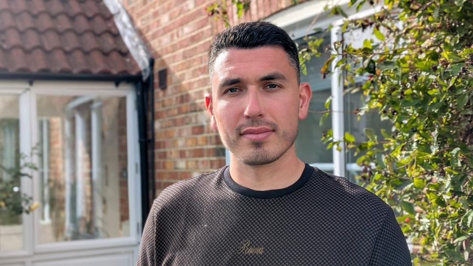 Mike, a man with short, dark hair and wearing a dark grey top, stands in the sunshine outside his house