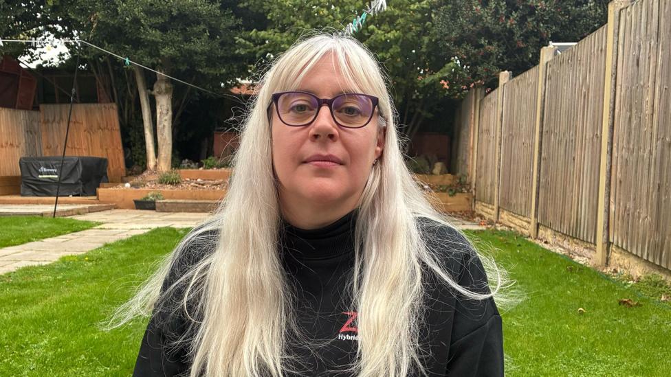 Rose-Marie, a woman with long, blonde hair and dark-framed glasses, is pictured in a garden.