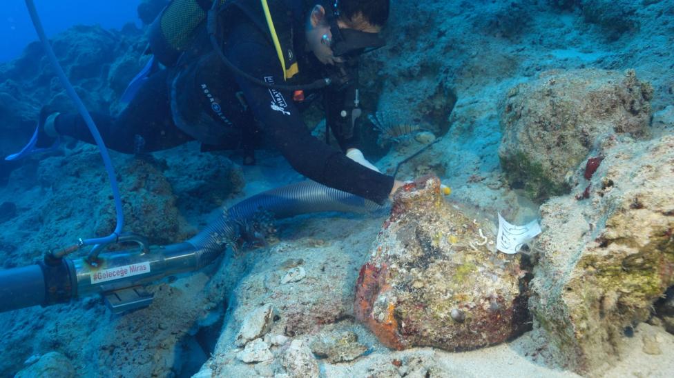 A diver on the top left hand side of the picture is working underwater to uncover an amphora. They are wearing a black wet suit with some white writing on it and a yellow stripe. They have black goggles. A long tube, like the sort you find on a vacuum cleaner, is being used to remove the sand that has been covering the ship's cargo. In the foreground and slightly to the right is an amphora. It is mostly covered in a sort of multicoloured rocky crust and it is sitting on a small ledge but some patches of the reddish clay are visible. There is a piece of paper with some numbers and letters written on it attached to the pot. To the right and behind is a small ridge of rocks.