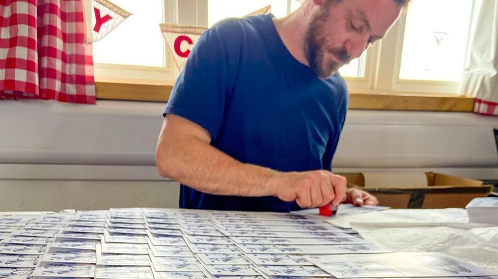 George Clarke is pictured stamping postcards at Port Lockroy, with dozens spread out across the table in front of him. He’s wearing a blue top and has brown facial hair.