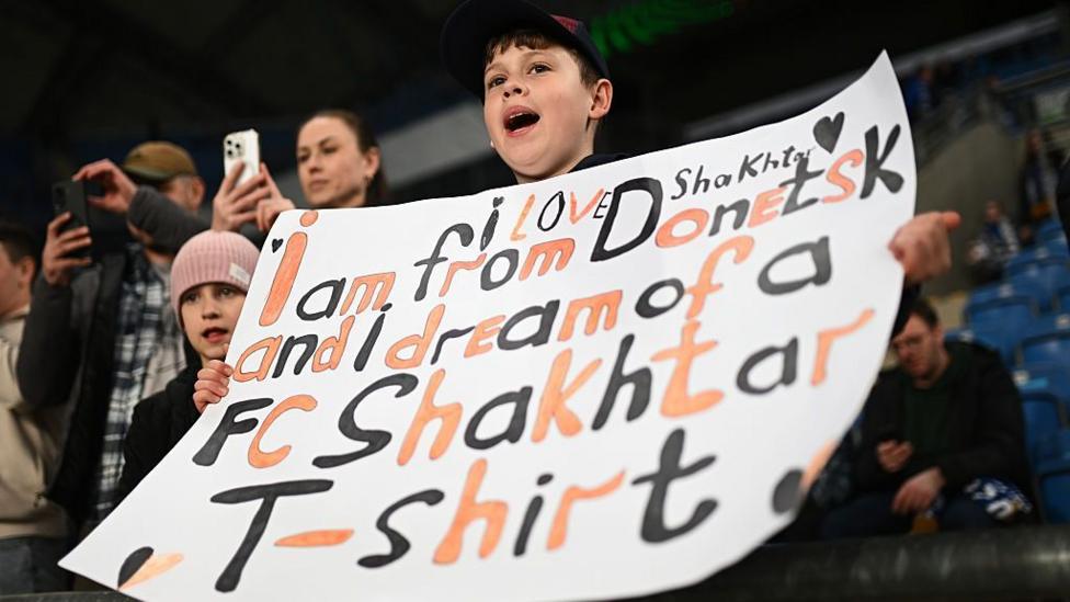 A young fan of Shakhtar Donetsk holds up a sign which reads "I love Shakhtar, I am from Donetsk and I dream of a FC Shakhtar Donetsk T-shirt" before a Conference League 2025-26 match against Lech Poznan