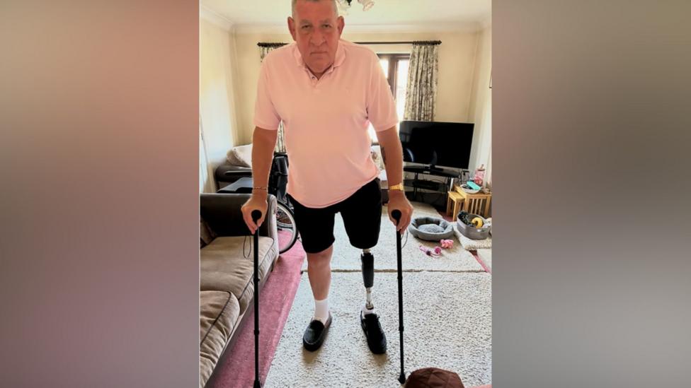 Nigel Ward stands in a living room using two walking sticks and a prosthetic leg. A wheelchair, sofa, television and pet bed can be seen in the background.