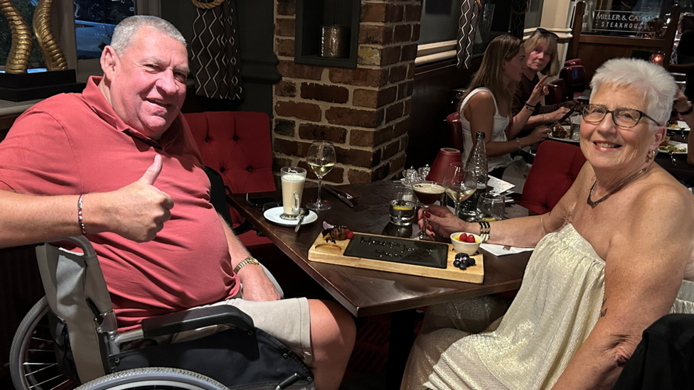 "A wheelchair user sits at a restaurant table beside another diner. The table holds drinks and dessert dishes, while other customers can be seen seated in the background."