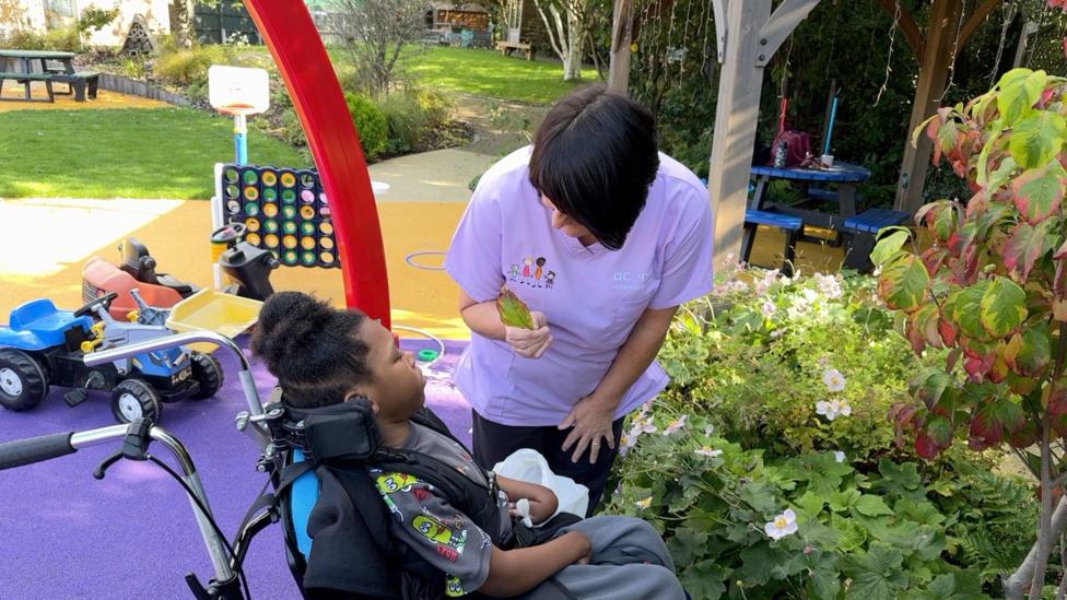 A young girl sits in a wheelchair outside a hospice, with a hospice worker in a lilac tunic standing next to her