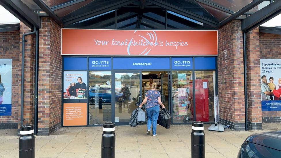 The exterior of an Acorns charity shop with orange signage. A woman can be seen walking through the door carrying black bin bags