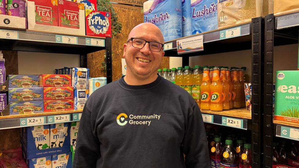A man with a bald head and glasses is wearing a black sweatshirt which says "Community Grocery" He is standing in front of shelves with food and household items