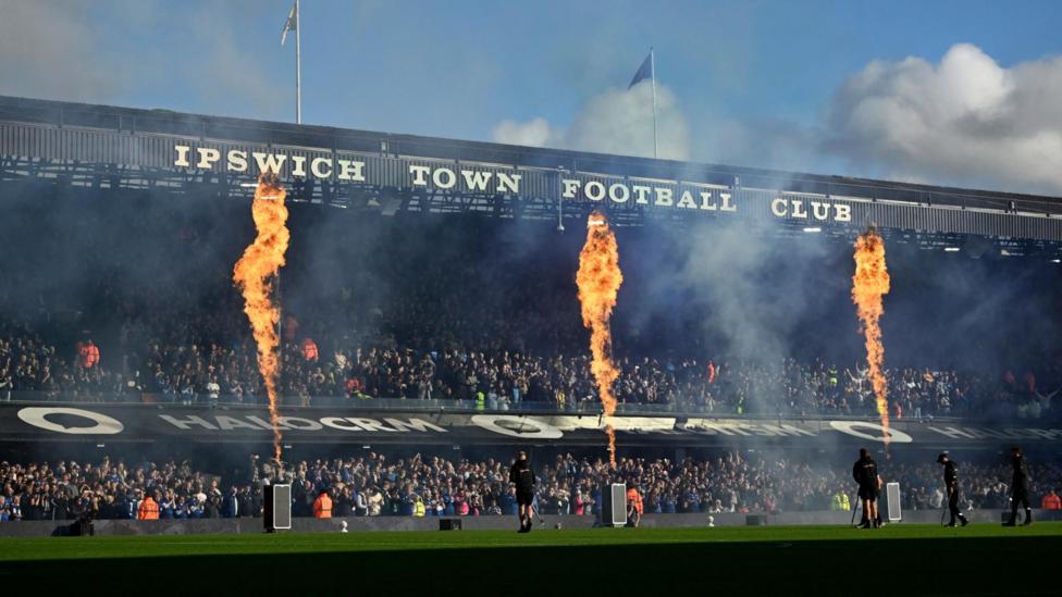 A two-tiered stand at a football ground. It has "IPSWICH TOWN FOOTBALL CLUB" in white letters on the front of the roof. Between the upper and lower tiers is a sloping roof over executive boxes which has advertising on it. There are three flames rising from devices on the pitch that reach the roof of the stand. The stand is full of supporters.