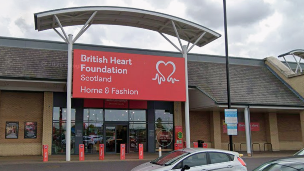 British Heart Foundation shop in a retail park. White writing on a red sign on top of the entrance reads British Heart Foundation Scotland Home & Fashion. Cars are parked in a car park outside.