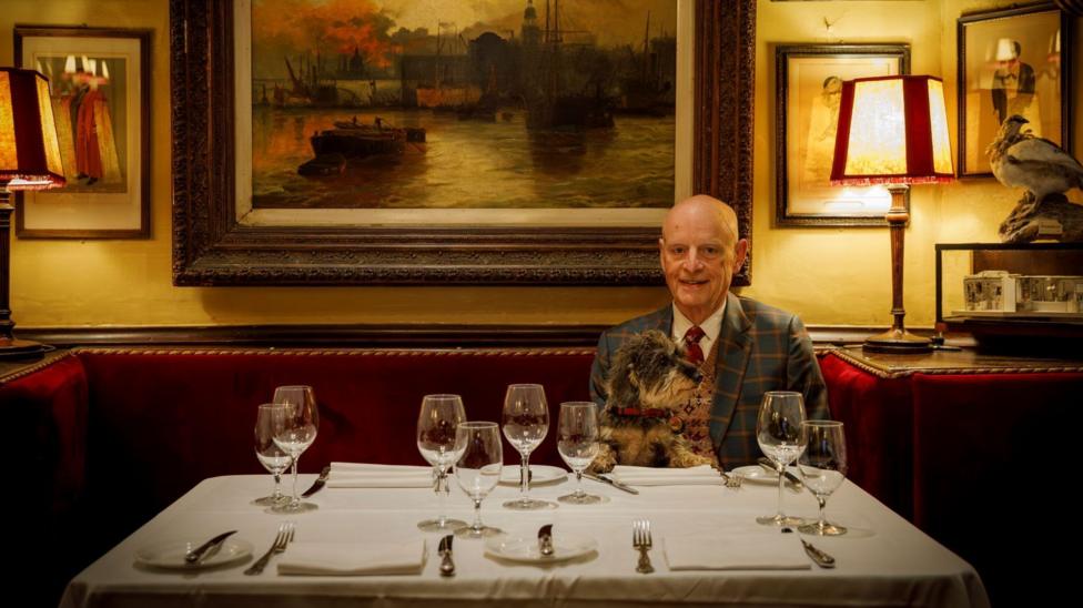 A balding man sits on a red sofa at a table with a white table cloth and wine glasses. He sits in front of a painting of ships.