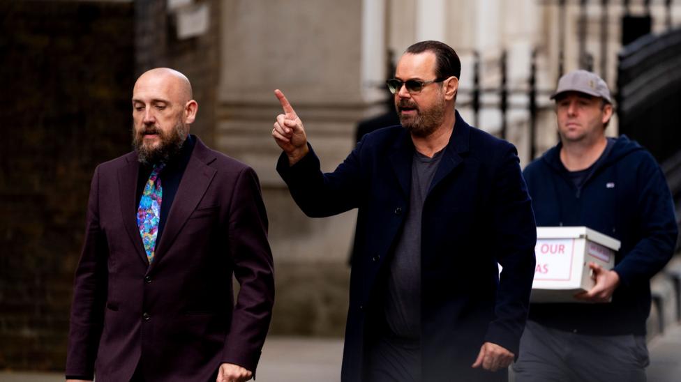 A man, Danny Dyer, walking down the street and pointing in front of him. He is with another man, Kevin McKenna, in a burgundy suit, and a third man is behind them carrying a white box.