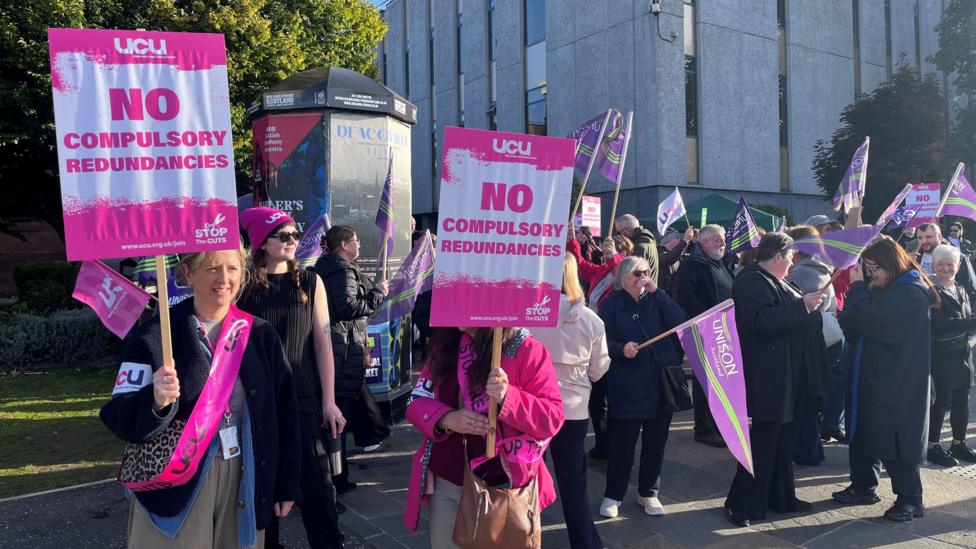 Protestors standing outside a university campus, with many waving placards or flags. The placards are in pink or purple and say things like NO COMPULSORY REDUNDANCIES in large capital letters.