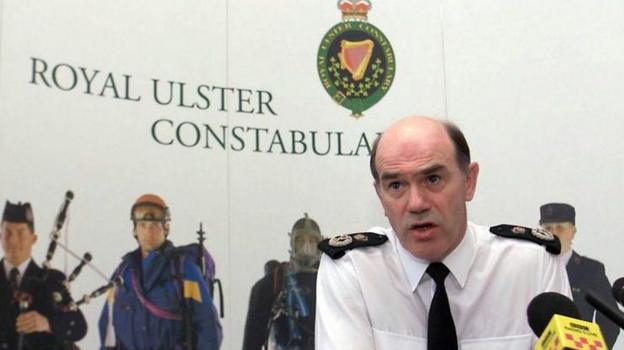 An archive image of Sir Ronnie Flanagan mid-speech at the launch of the RUC Annual Report in 2000. He has receding short, black hair and is wearing a white RUC uniform shirt with black and gold epaulettes and a black tie. A poster with a green and white RUC crest and five people dressed in different uniforms and suits is in the background.