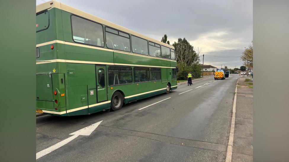 A green double-decker bus parked on the side of the road. A police officer can be seen in the distance and a flashing police van. 