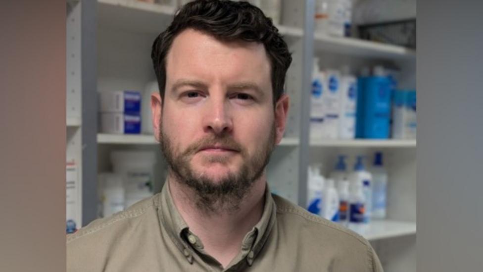 A man with dark hair and a short beard looks into the camera. He is wearing a beige top and there are shelves of medication behind him.