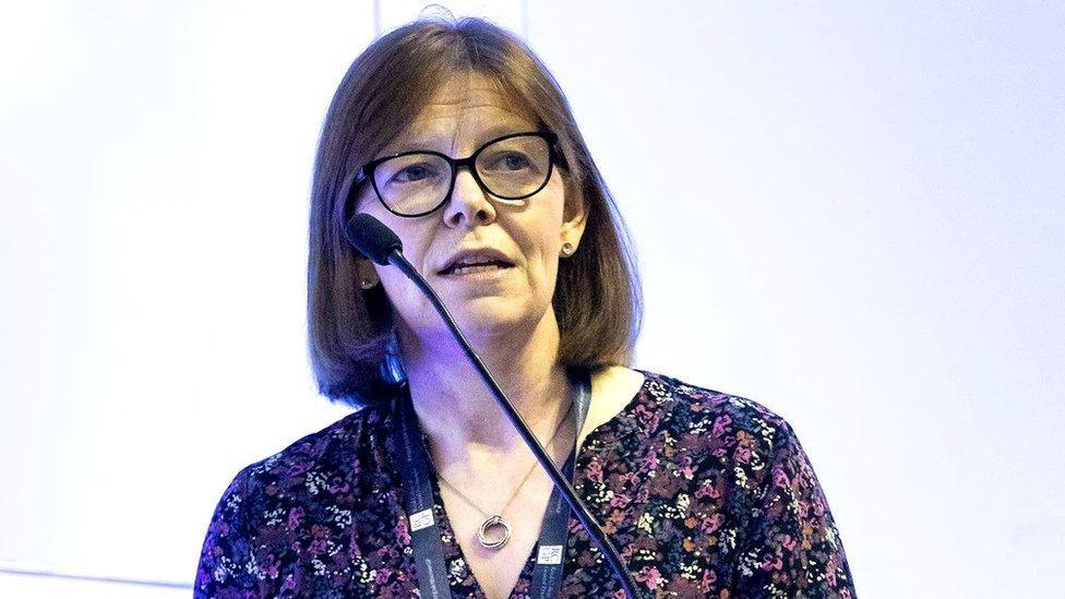 A close-up image of Dr Ursula Mason speaking at a podium with a large mic. She has shoulder-length brown hair and is speaking into a microphone. She is wearing a pair of dark, circular glasses, a patterned shirt and she is standing in front of a light wall looking out into the crowd.
