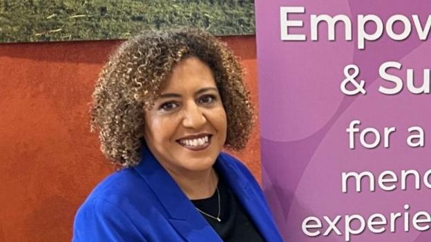 A woman with curly bob and wearing a royal blue jacket and a navy top stands beside a board encouraging women to become empowered.