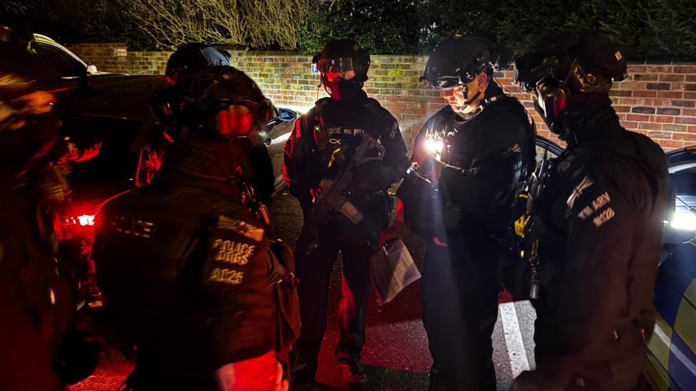Armed police are gathered for a briefing ahead of a raid. You can see officers wearing helmets and holding weapons, with parked cars in the background, including a patrol car.