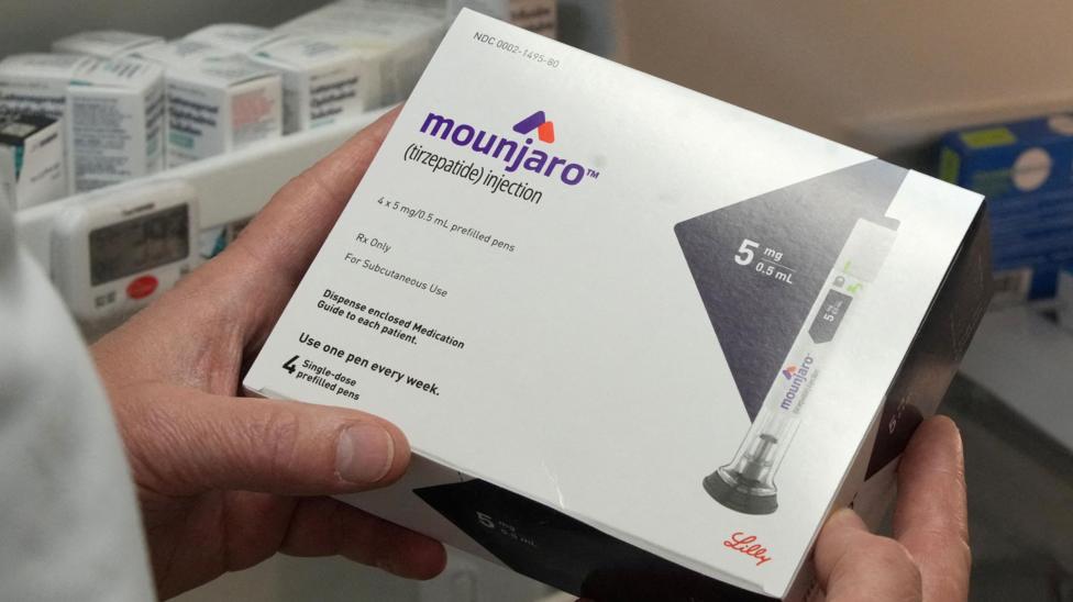 A pharmacist holding a box of Mounjaro. Other medicines are aranged in the background.