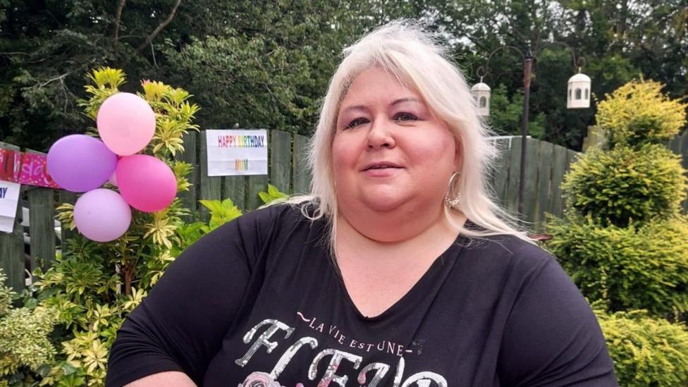 A woman with blonde hair and a black top in a garden, with balloons and a happy birthday sign in the background.