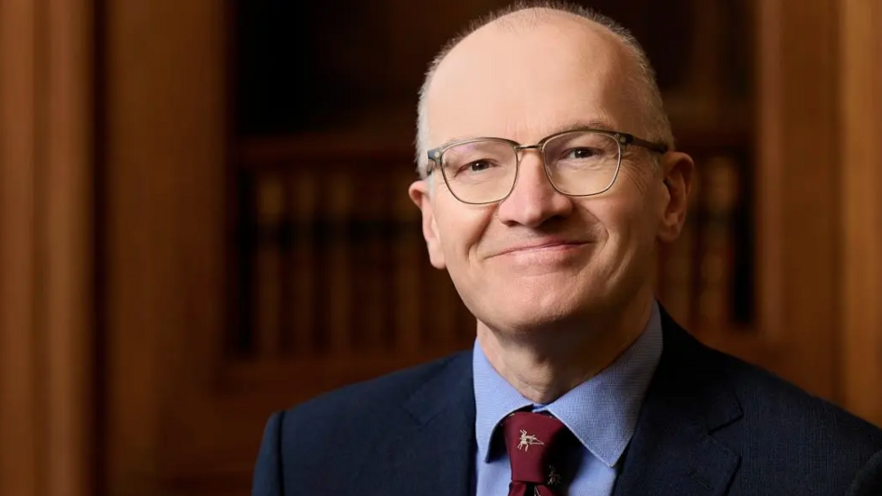Dr Mark Strachan, a man with short grey hair and glasses, pictured in front of a bookcase. He is wearing a darks blue suit, light blue shirt and a burgundy tie.