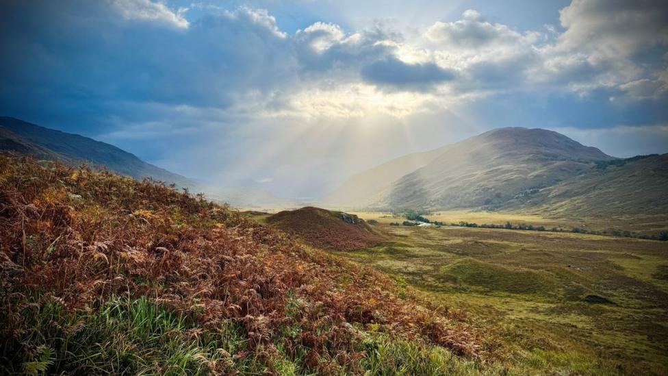 A valley surrounded by hills and mountains with the sun shining behind the clouds