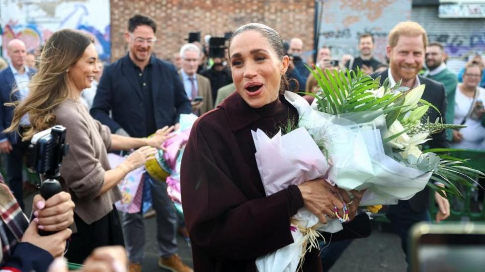 Meghan smiles broadly in the centre of the picture holding her flowers as Harry can be seen in the background out of focus but grinning
