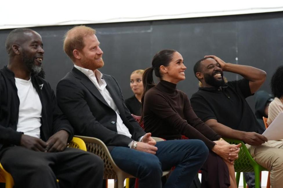 The Duke sits relaxed in the hall alongside the Duchess who has her hands clasped together and is grinning in her seat